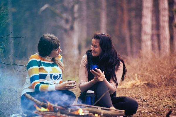 two women sitting in forest by fire smiling and talking